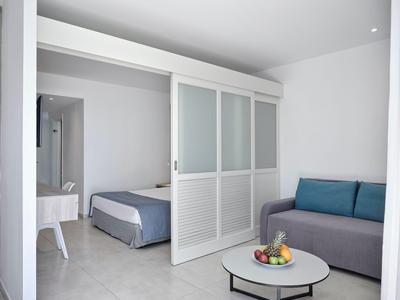 Hell eingerichtetes Hotelzimmer mit Schlafbereich, Sofa und Obst auf einem runden Tisch.