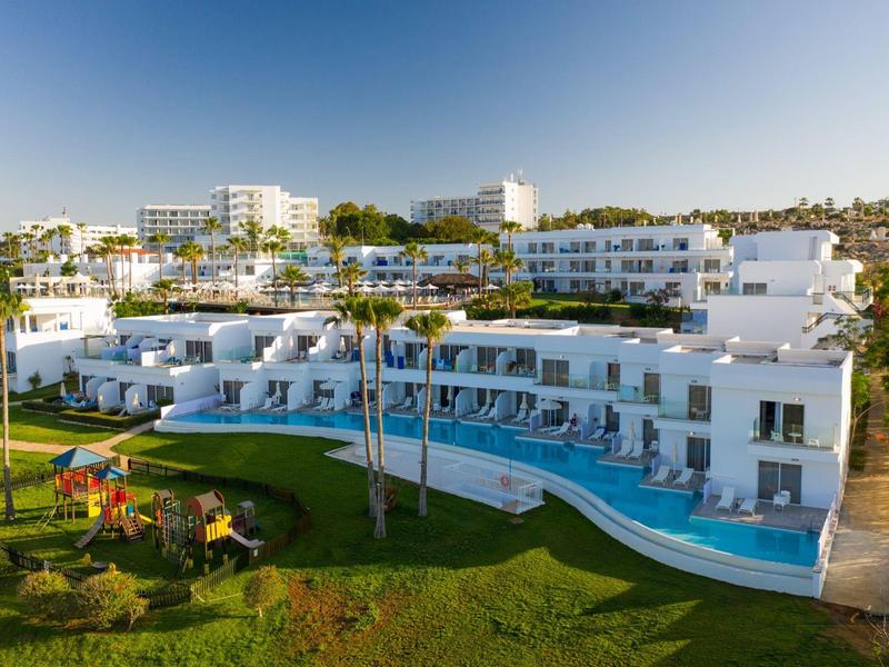 Moderne weiße Hotelanlage mit Pool und grüner Gartenanlage unter klarem Himmel.