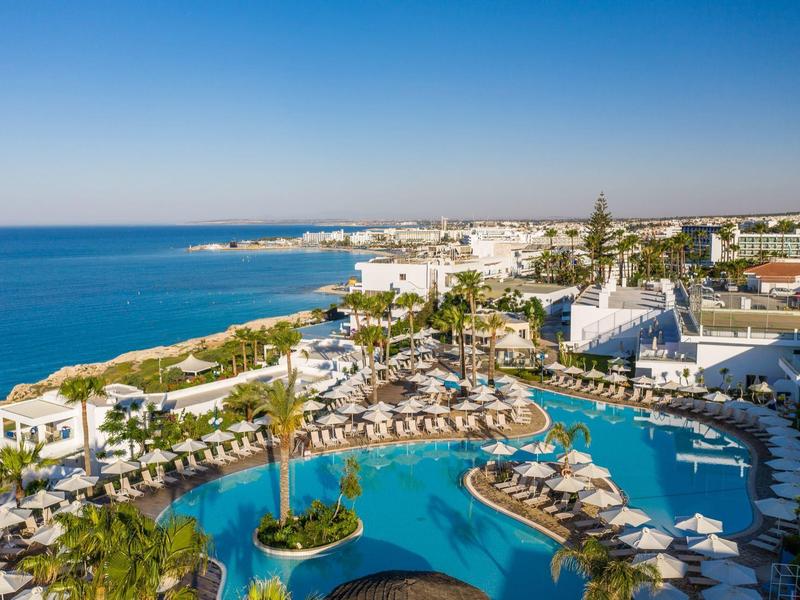 Blick auf ein großes Resort mit mehreren Pools, Liegestühlen und Palmen am Meer unter klarem Himmel.