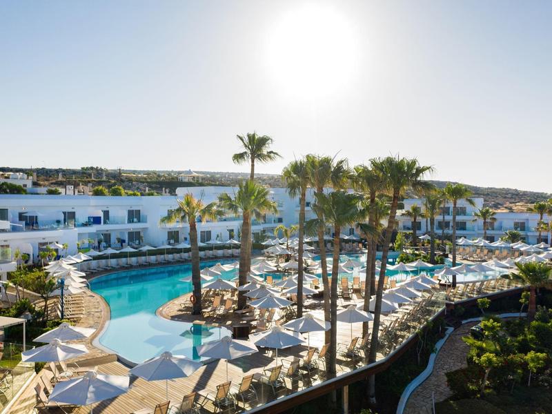 Großer Hotelpool mit Palmen, Liegestühlen und weißen Gebäuden bei sonnigem Himmel.