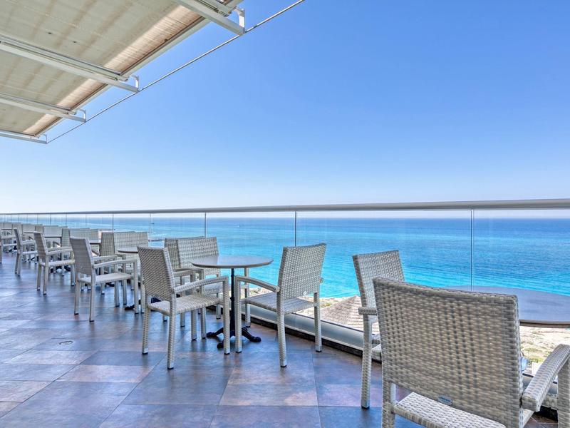 Terrassenbereich mit modernen weißen Tischen und Stühlen, gläsernem Geländer und Blick aufs blaue Meer.