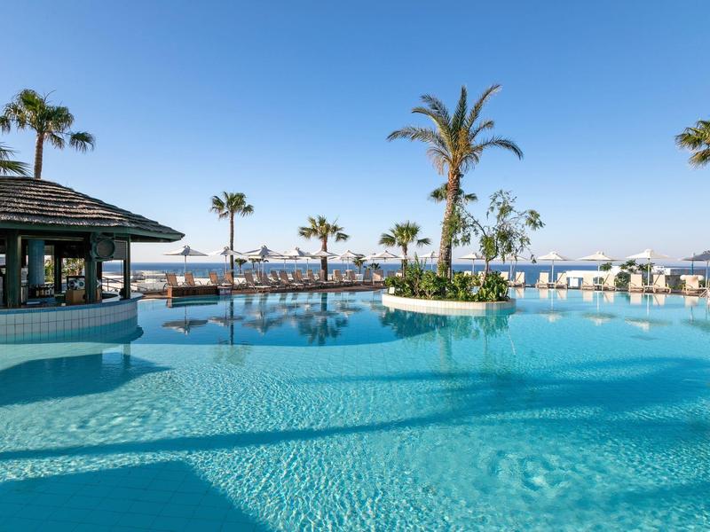 Großer blauer Pool mit Palmen, Sonnenschirmen und klarem Himmel am Meer im Hintergrund.