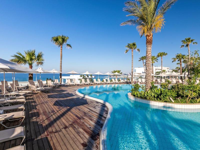 Moderner Poolbereich mit Palmen, Liegestühlen, Holzdeck und Blick auf das blaue Meer.