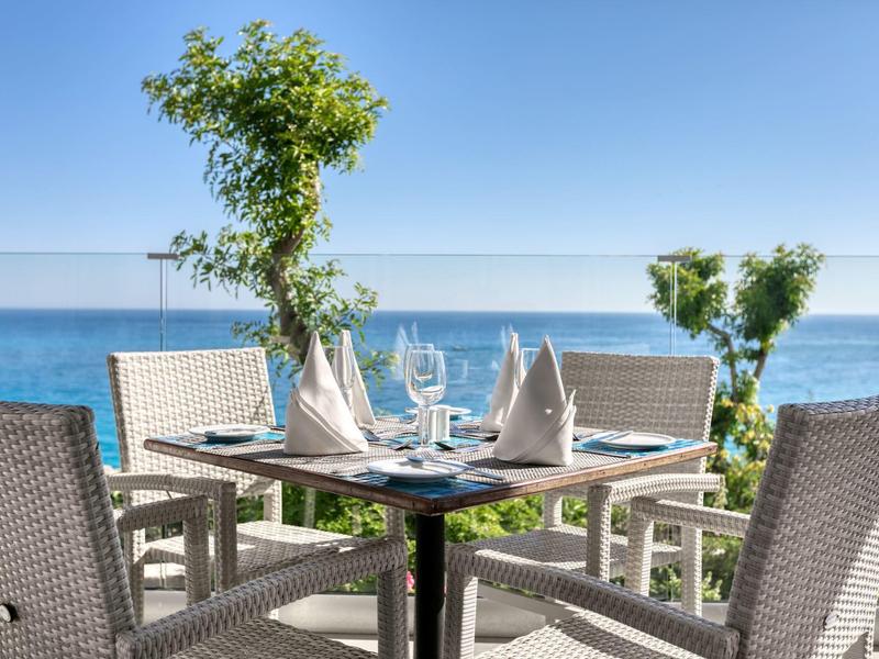 Vier geflochtene Stühle um runden Tisch mit eingedeckten Gläsern und Servietten, Meer im Hintergrund.