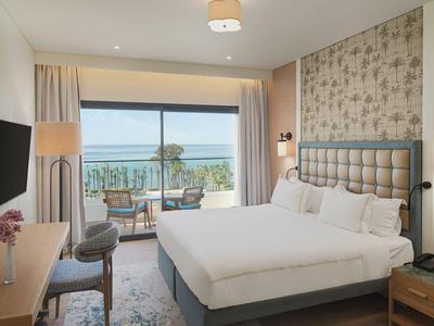 Habitación de hotel moderna con cama doble, vista al mar y balcón con área de descanso.