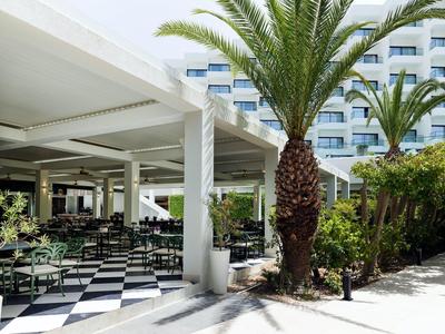 Außenbereich mit weißem Pergoladach, schwar-weiß kariertem Boden und Palmen vor einem Hotel.