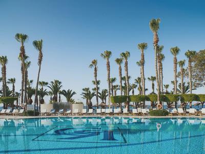 Großer Pool mit klarem Wasser, umgeben von hohen Palmen und weißen Liegestühlen unter blauem Himmel.