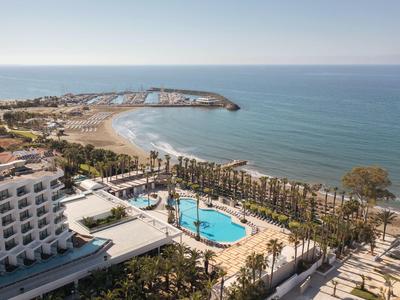Küstenhotel mit Pool, Palmen und Strand unter blauem Himmel am ruhigen Meerufer.