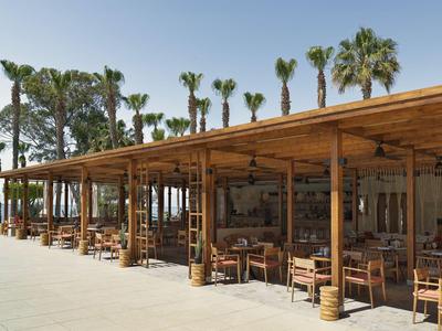 Restaurante al aire libre con techo de madera y palmeras junto al mar en un entorno soleado.