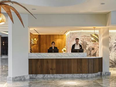 Moderner Empfangsbereich mit zwei Mitarbeiter*innen hinter hellem Tresen und Palmen-Dekor.