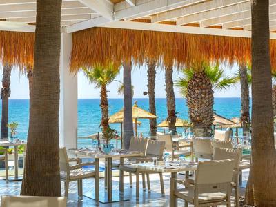 Offene Strandbar mit weißen Stühlen, Holztischen und Palmen vor blauem Meer und Himmel.