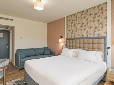 Modernes Schlafzimmer mit großem Bett, grauem Sofa, Holzmöbeln und floraler Wanddekoration.