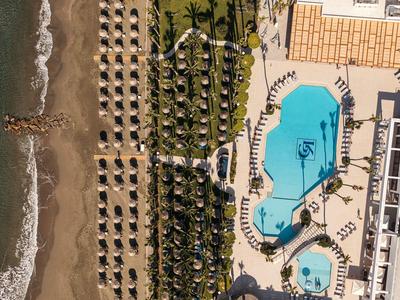 Strand mit Sonnenschirmen, Palmen und zwei blauen Pools neben einem großen Gebäude.