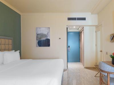 Helles Hotelzimmer mit großem weißen Bett, grün-blauer Wand, Kunstbild und Tisch mit Blumen.