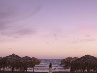 Frau am Strand mit Strohdächern, sanfter Wellen und rosa-violettem Himmel zum Sonnenuntergang.