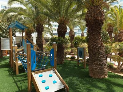 Kleiner Spielplatz mit Klettergerüst, Rutsche und Palmen auf grünem Kunstrasen im Freien.