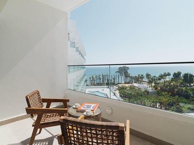Balkon mit zwei Stühlen und Tisch, Blick auf Palmen und Meer bei klarem Himmel.