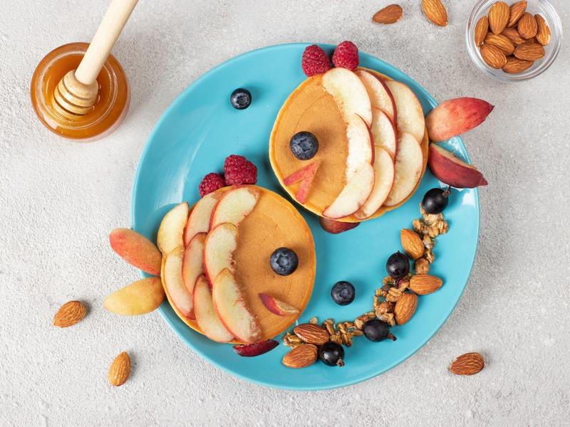 Zwei Pfannkuchenfische auf blauem Teller mit Apfel, Beeren, Mandeln, Honig und Nüssen arrangiert.