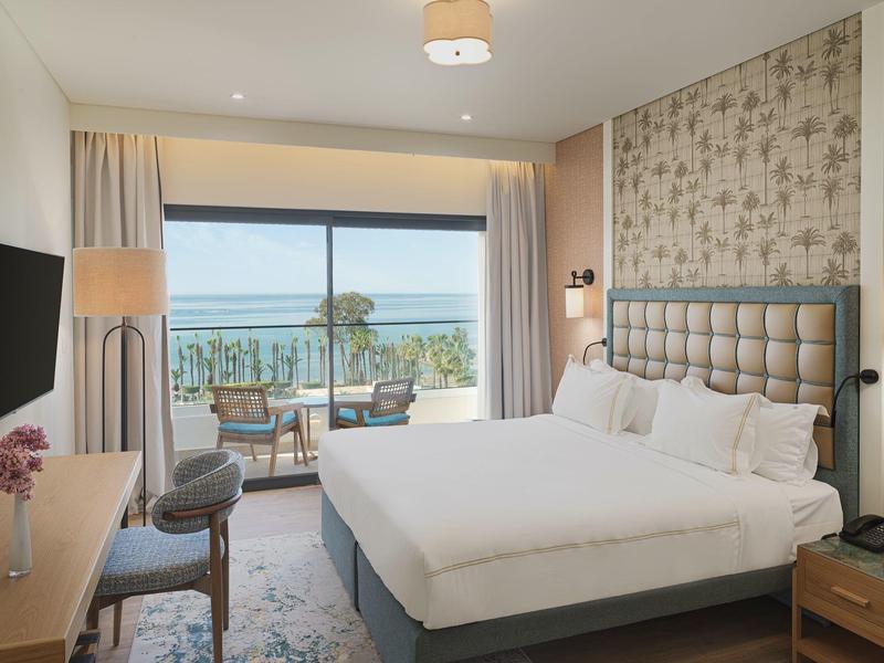 Modernes Hotelzimmer mit Doppelbett, großem Fenster und Balkon mit Meerblick. Helle, warme Farben.