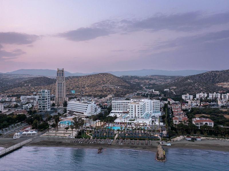 Stadt mit hohen Gebäuden, Hügeln im Hintergrund und Strand mit Promenade im Vordergrund bei Abendlicht.