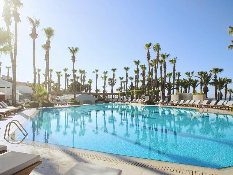 Großer Pool mit klarem blauen Wasser, umgeben von Palmen und Liegestühlen unter blauem Himmel.
