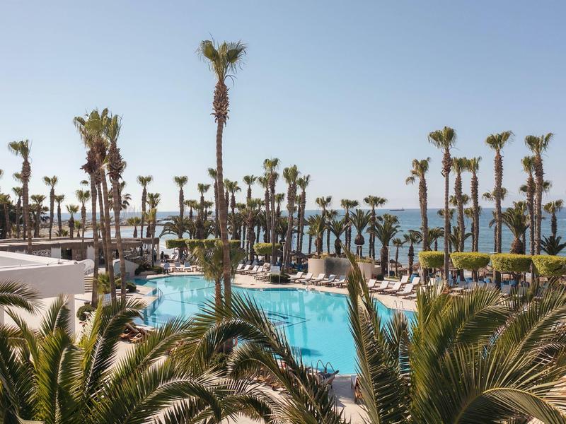 Ein blauer Pool umgeben von hohen Palmen und grünen Pflanzen unter klarem Himmel am Meer.