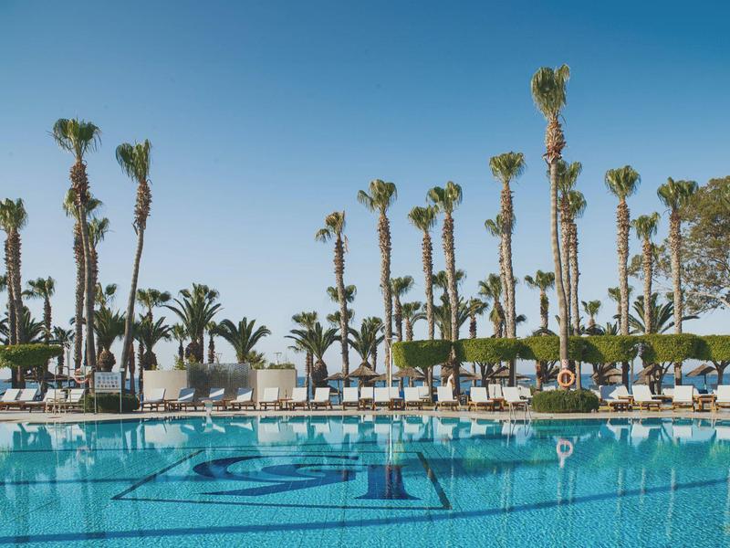 Großer Pool mit klarem Wasser, umgeben von hohen Palmen und weißen Liegestühlen unter blauem Himmel.