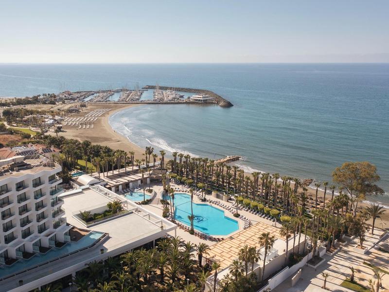 Küstenhotel mit Pool, Palmen und Strand unter blauem Himmel am ruhigen Meerufer.