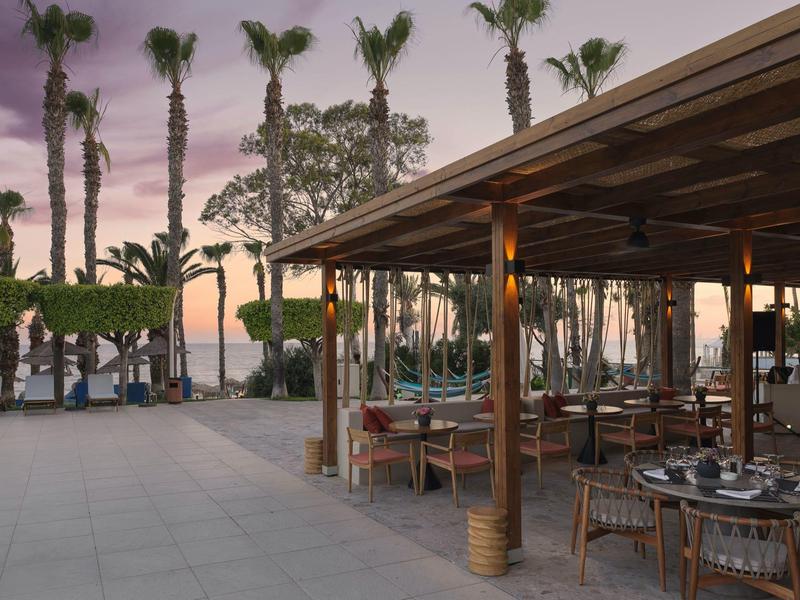 Terrasse mit gedeckten Tischen, Palmen und Sonnenuntergang am Meer im Hintergrund.