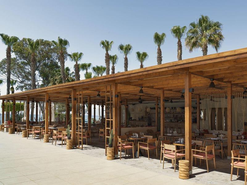 Offenes Restaurant mit Holzdach, Stühlen und Tischen neben Palmen und blauem Himmel.
