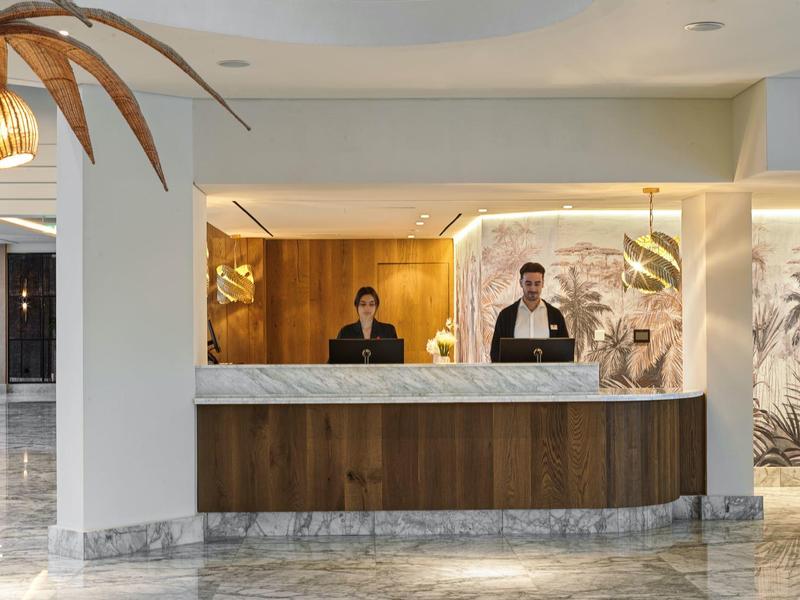 Moderner Empfangsbereich mit zwei Mitarbeiter*innen hinter hellem Tresen und Palmen-Dekor.