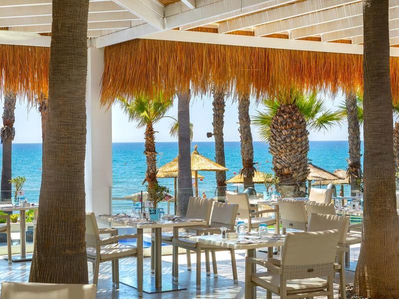 Offene Strandbar mit weißen Stühlen, Holztischen und Palmen vor blauem Meer und Himmel.