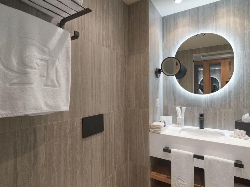 Modernes Badezimmer mit hellen Holzwänden, weißem Waschbecken, rundem Spiegel und Handtüchern.