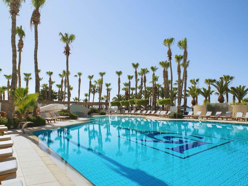 Großer rechteckiger Pool mit klar blauem Wasser, umgeben von Liegestühlen und Palmen unter blauem Himmel.