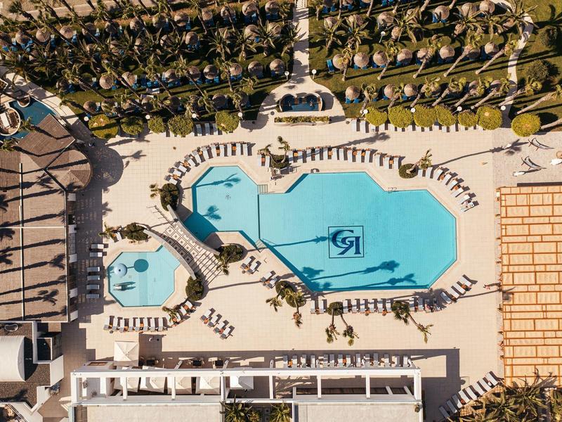 Vogelperspektive auf eine große, geformte blaue Poolanlage mit umliegenden Liegestühlen und Palmen.