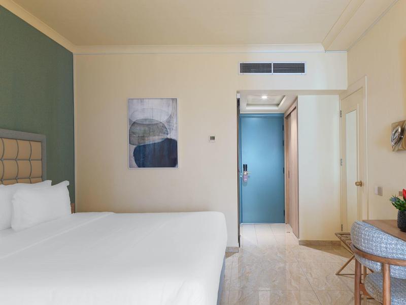 Helles Hotelzimmer mit großem weißen Bett, grün-blauer Wand, Kunstbild und Tisch mit Blumen.