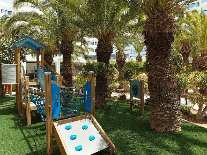 Kleiner Spielplatz mit Klettergerüst, Rutsche und Palmen auf grünem Kunstrasen im Freien.