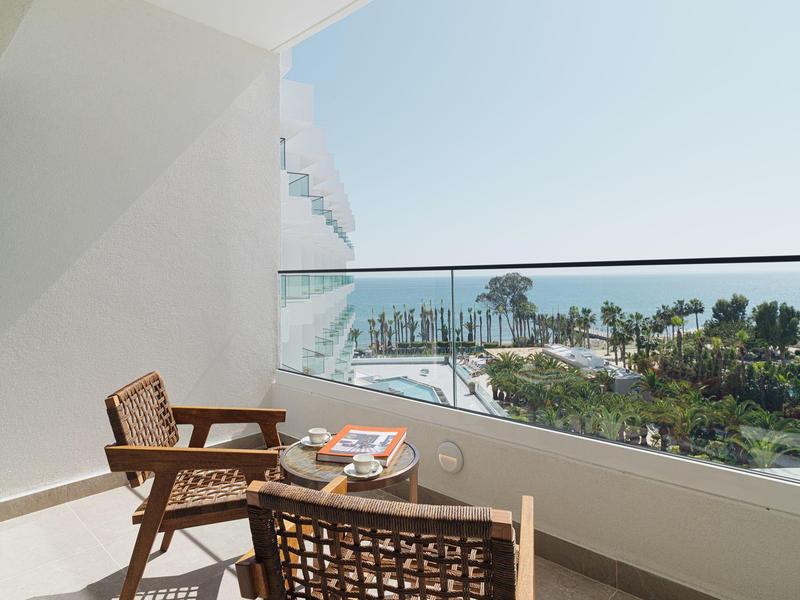 Balkon mit zwei Stühlen und Tisch, Blick auf Palmen und Meer bei klarem Himmel.