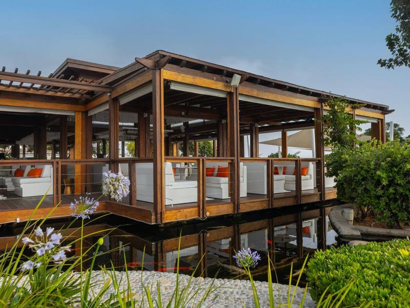 Moderner Pavillon aus Holz mit Glaswänden, umgeben von Wasser, Pflanzen und blauen Himmel.