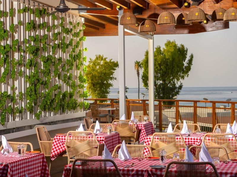 Offenes Restaurant mit rot-weiß karierten Tischtüchern, Holztischen, grünen Pflanzen und Meerblick.