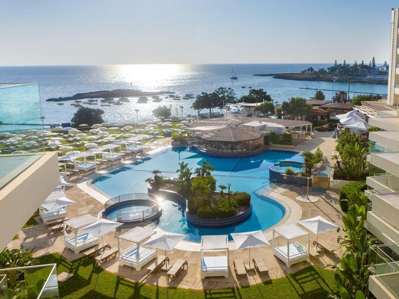 Blick auf Hotelpool, Liegestühle und Meer bei Sonnenuntergang, mit Gebäuden im Hintergrund.