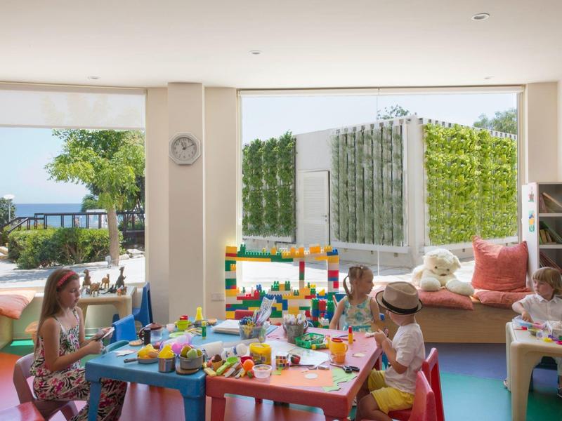 Kinder spielen im hellen Raum mit buntem Spielzeug an einem blauen Tisch, draußen grünt es.