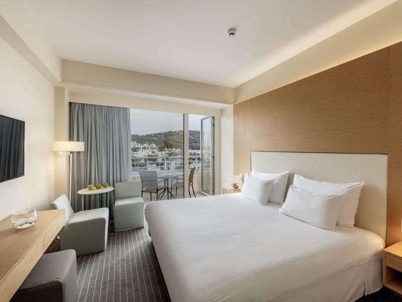 Modernes Hotelzimmer mit großem Bett, Fernseher, Schreibtisch und Balkon mit Blick auf Hafen.