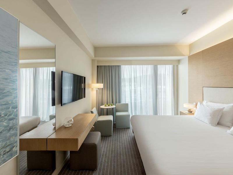 Helles Hotelzimmer mit großem Bett, Flachbildfernseher, Vorhängen und modernem Möbelstil.