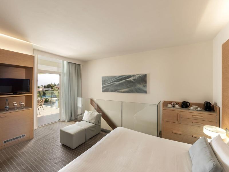 Helles Hotelzimmer mit Bett, Holzkommode, TV, Balkon und hellem Teppichboden.