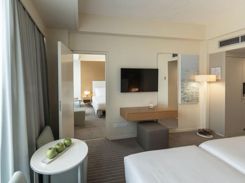 Modernes, helles Hotelzimmer mit Doppelbett, TV, Schreibtisch, grauen Vorhängen und Blick ins angrenzende Zimmer.