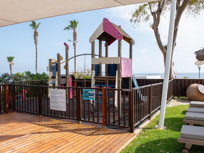 Plac zabaw dla dzieci z drabinką i zjeżdżalnią na ogrodzonym terenie obok tarasu i roślin.