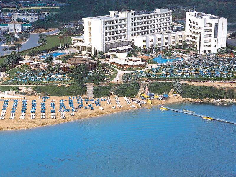 Großes weißes Hotel am Strand mit blauem Meer, Liegestühlen und grünen Bäumen im Hintergrund.