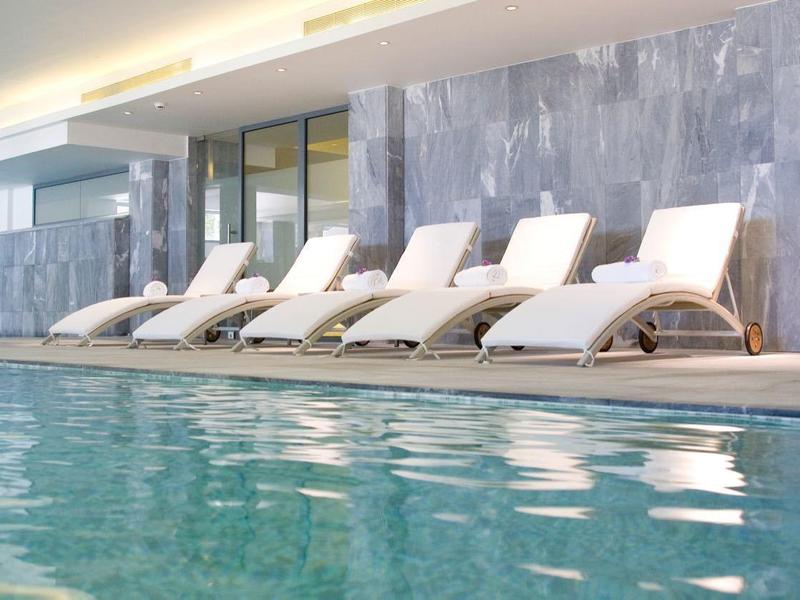 Ein Hallenbad mit klarem Wasser, sieben weißen Liegen und grauen Fliesen an der Wand.