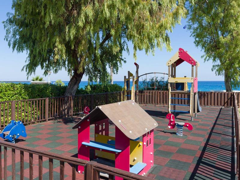 Kinderspielplatz mit buntem Kletterhaus, Rutsche, Bäumen und Meer im Hintergrund unter blauem Himmel.